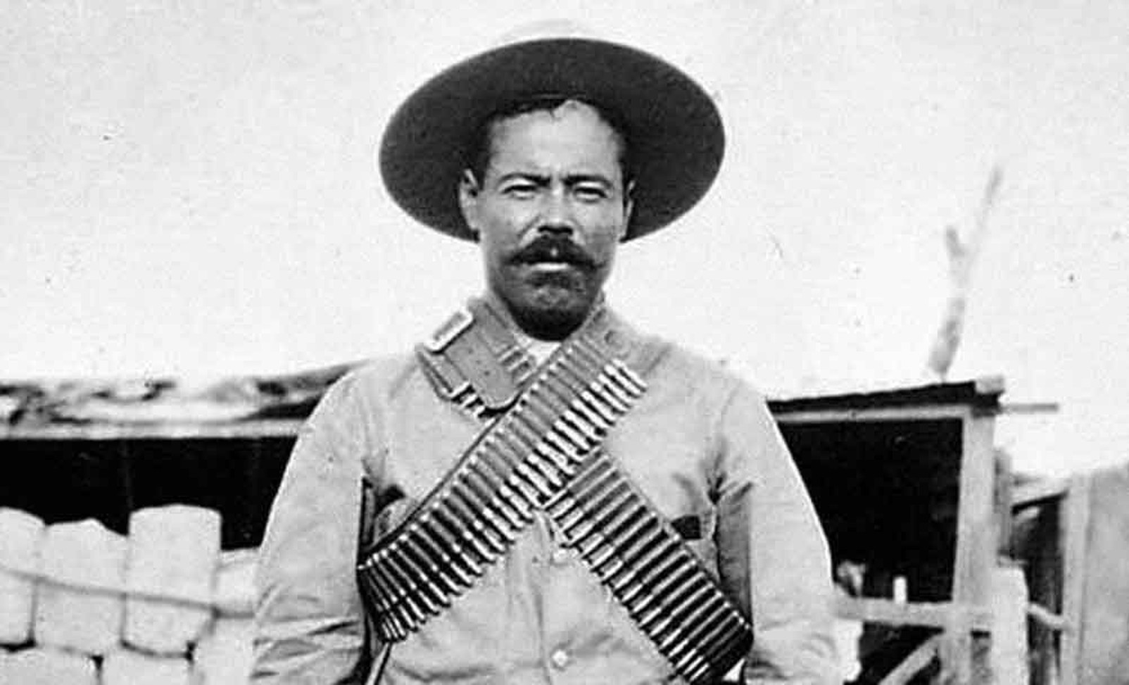 Archival photograph: Francisco “Pancho” Villa in a wide-brimmed hat and crossed bandoliers, standing in front of a plank wall