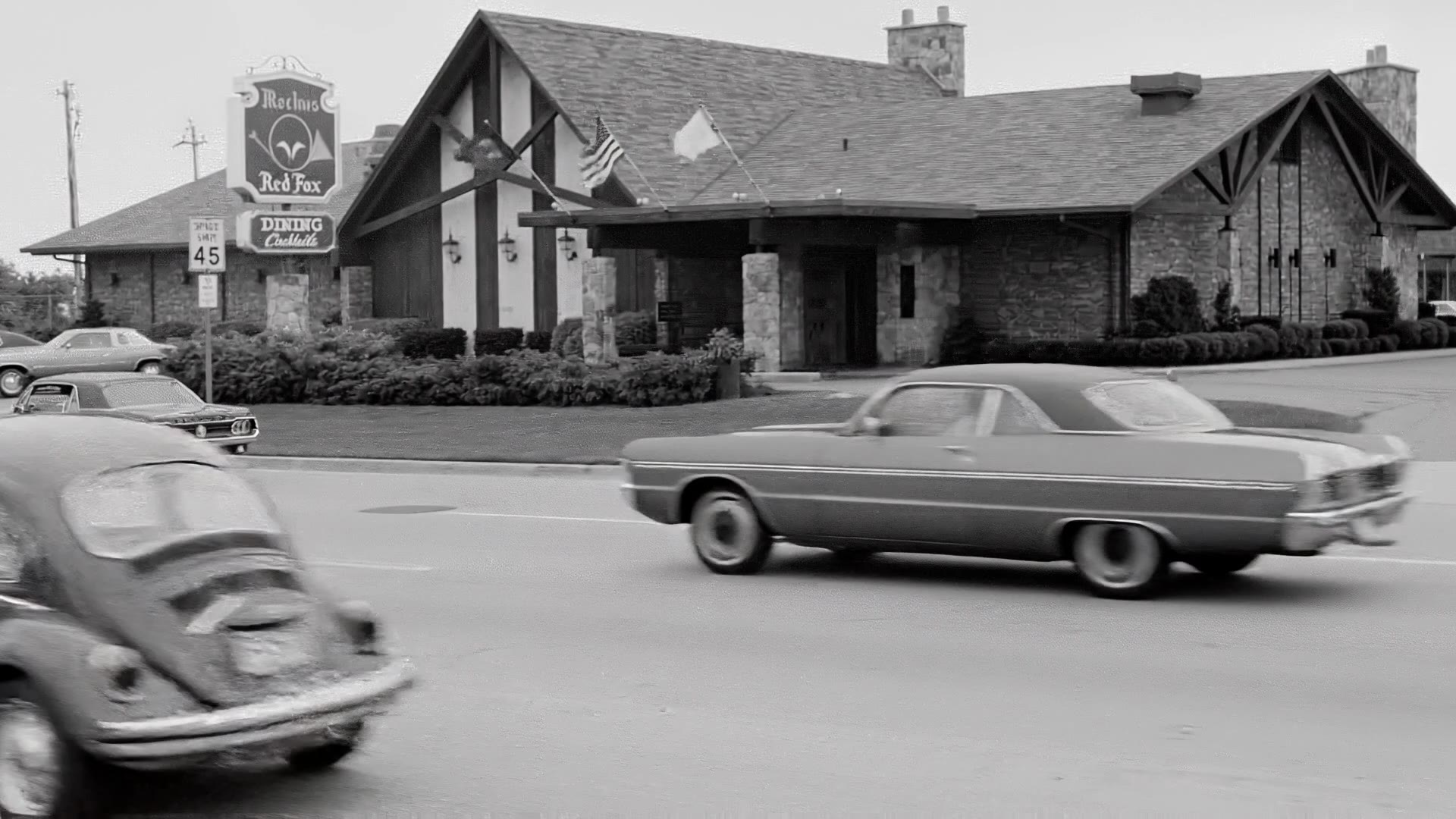Animated archival photograph: cars pass the Machus Red Fox steakhouse in suburban Detroit in black and white
