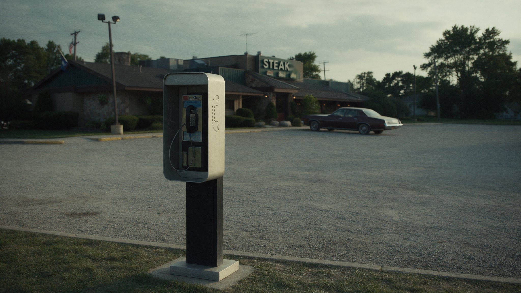A public payphone on a post at the edge of a gravel steakhouse parking lot, with a dark sedan parked near the building under an overcast sky